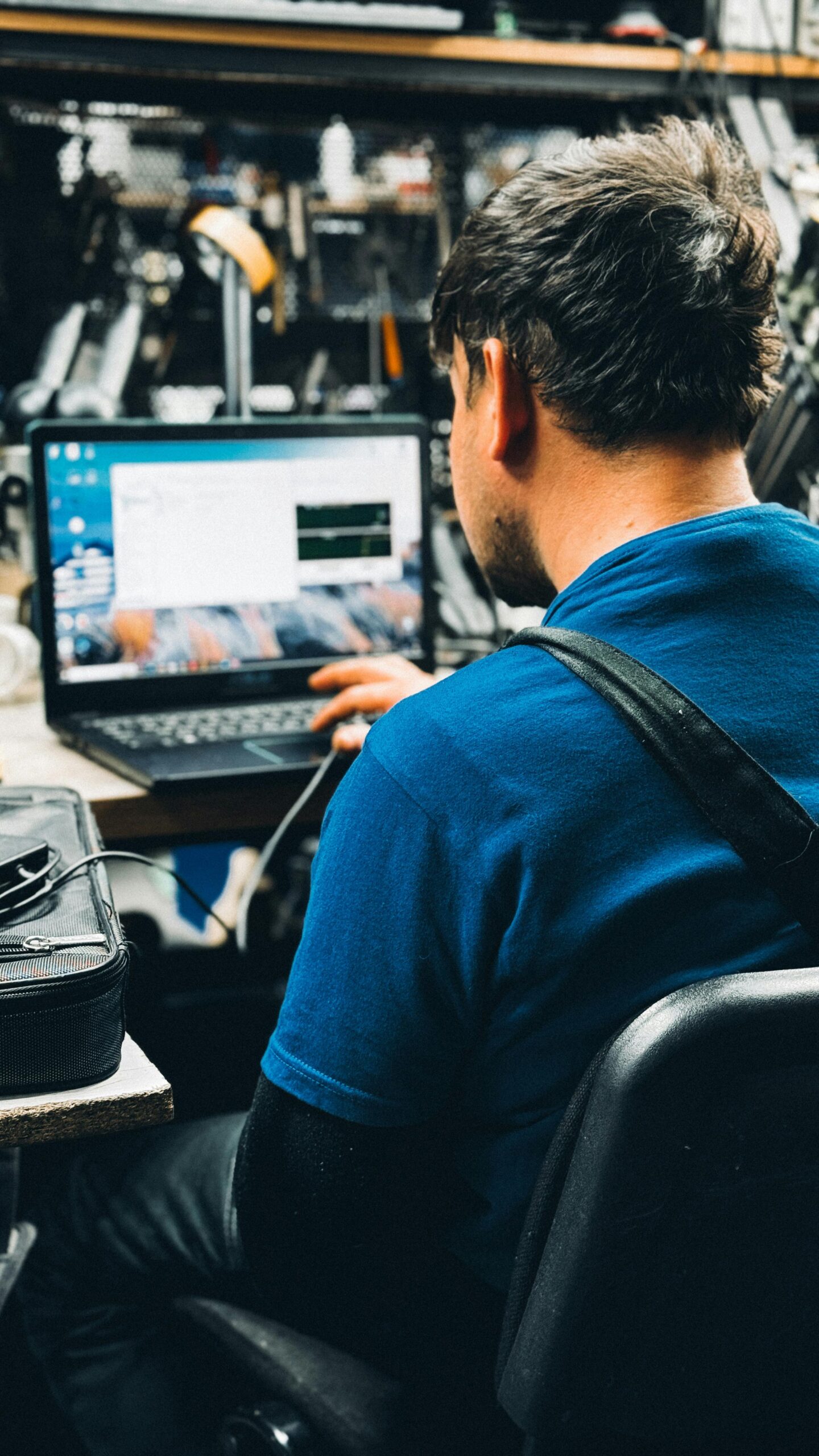 Technician in blue shirt working on a laptop in a busy workshop setting.